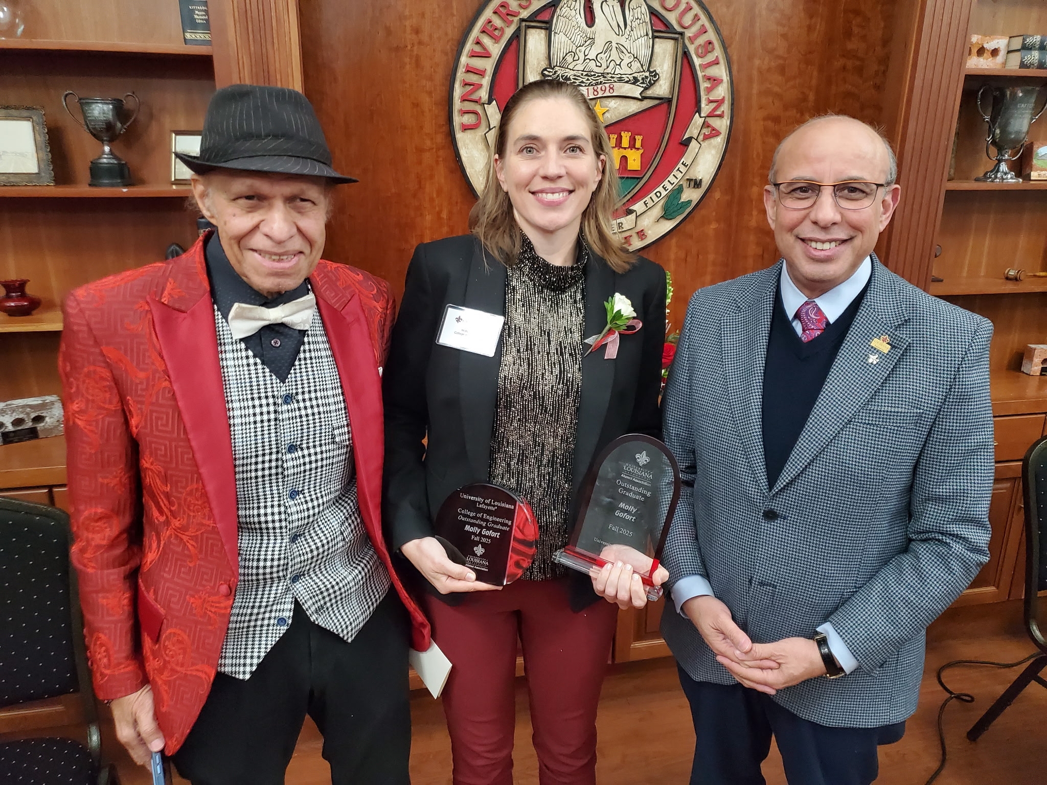 Picture of three people from left: Dr. Magdy Bayoumi, Department Head of Electrical and Computer Engineering, Ms. Molly Goforth hold her outstanding graduate awards and Dr. Ahmed Khattab, Dean of the College of Engineering.  All three are standing in front of a bookcase in the board room of the Alumni Center.